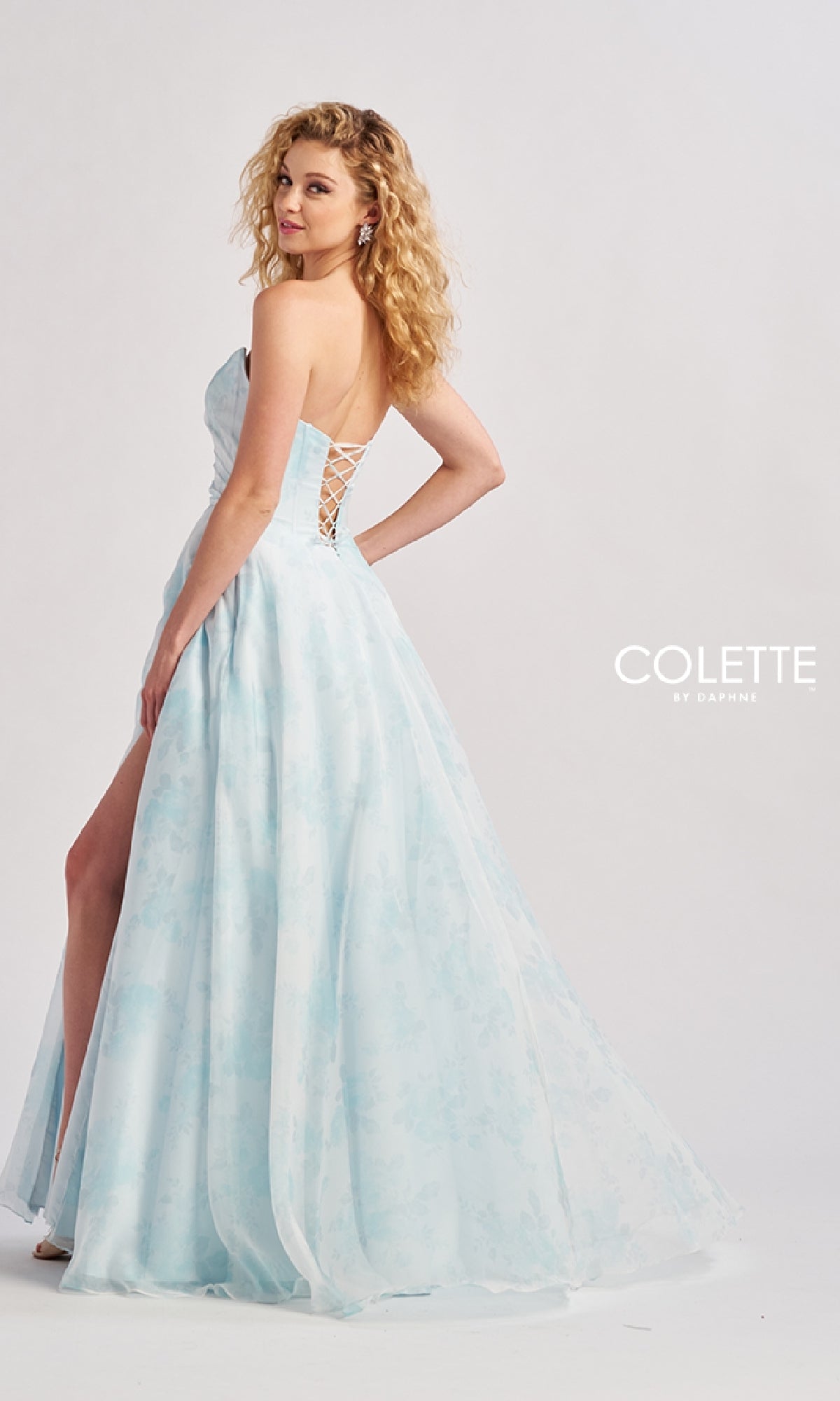 A woman poses in the Colette CL8635, a long light blue strapless prom ball gown featuring a lace-up back and high slit, standing against a white background. This elegant pastel dress is perfect for prom.